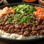 A bowl of ground beef bulgogi garnished with sesame seeds and green onions, served with steamed rice and lettuce wraps on a rustic wooden table.