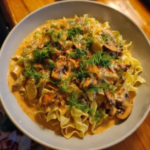 A close-up of creamy Vegan Mushroom Stroganoff with noodles, fresh dill, and paprika in a gray bowl on a wooden table.