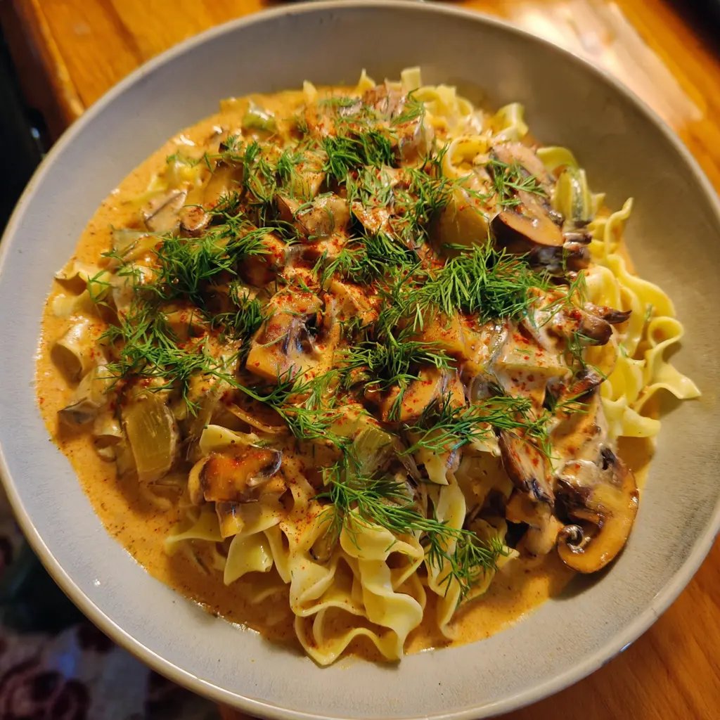 A close-up of creamy Vegan Mushroom Stroganoff with noodles, fresh dill, and paprika in a gray bowl on a wooden table.