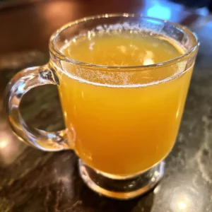 Clear glass mug with a vibrant orange-yellow beverage, potentially a refreshing Natural Mounjaro Recipe, on a dark reflective surface.