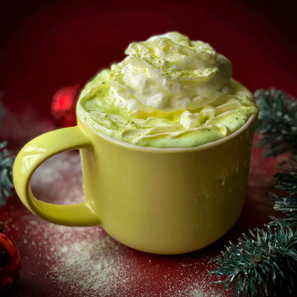 Festive green Grinch hot chocolate recipe in a lime green mug with whipped cream and green powder.