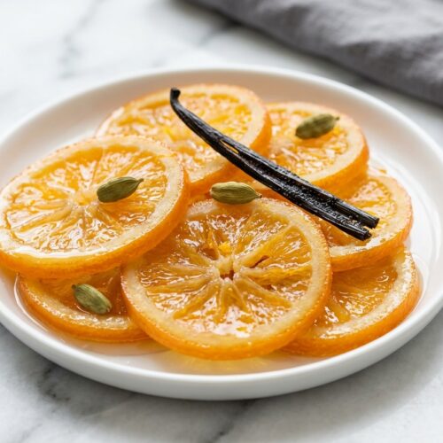 Translucent candied orange slices infused with vanilla bean and cardamom on a minimalist ceramic plate over white marble.