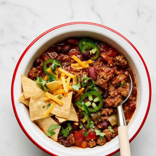 Ground beef chili recipe in a minimalist ceramic bowl on a white marble surface, showing tender beef and rich cocoa powder depth.