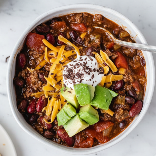 Rich venison chili recipe in a minimalist ceramic bowl on white marble, glossy mole finish with dark chocolate.