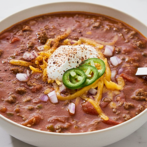 Flat lay of a texas roadhouse chili recipe in a minimalist ceramic bowl on white marble, highlighting the grainy masa harina texture and dark espresso hue.