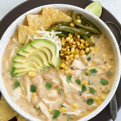 White chicken chili recipe in a minimalist ceramic vessel on a white marble surface with blistered poblano and charred corn.