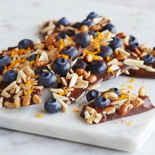 High-angle shot of blueberry chocolate bark on white marble, featuring bright orange zest and a hint of smoked paprika.