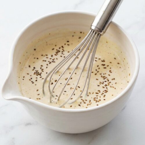 Creamy Coleslaw Dressing in a minimalist ceramic bowl on white marble, featuring toasted celery seeds and dill pickle brine.