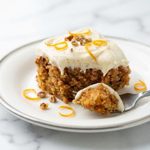 Gluten-Free Carrot Cake with speckled browned butter cream cheese frosting and orange zest on a white marble surface.