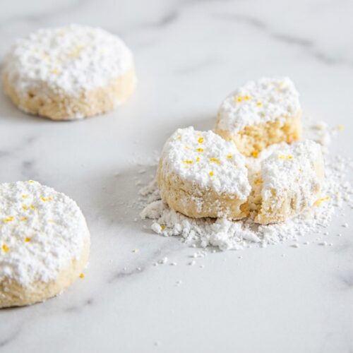 Meyer Lemon Meltaways stacked on a minimalist ceramic plate, dusted with fine citrus zest powder on a white marble surface.