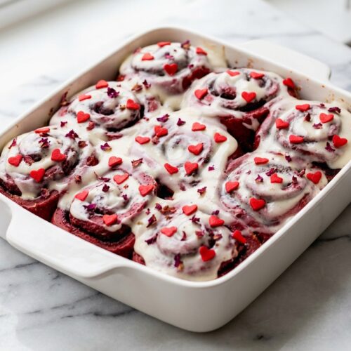 Heart-shaped Red Velvet Valentine's Day Cinnamon Rolls with rose water cream cheese frosting on a white marble surface.