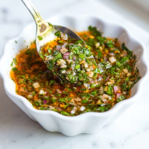 Vibrant Chimichurri Sauce in a minimalist ceramic bowl on white marble, showing the rich 'bloomed' spice oil infusion.