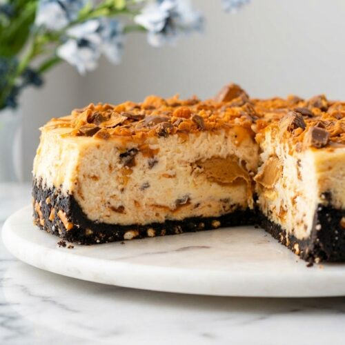 Creamy Butterfinger Cheesecake slice featuring a Salted Pretzel-Oreo crust on white marble with natural window lighting.