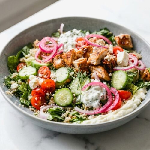 Greek Chicken Bowls served over double-lemon whipped feta and sumac pickled onions in a minimalist bowl on white marble.