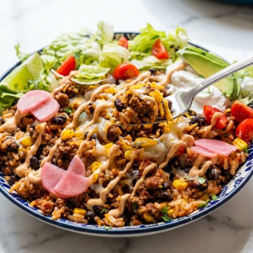 A vibrant Taco Rice Bowl in a minimalist ceramic vessel on a white marble surface, topped with chipotle-lime crema and pickled radishes.