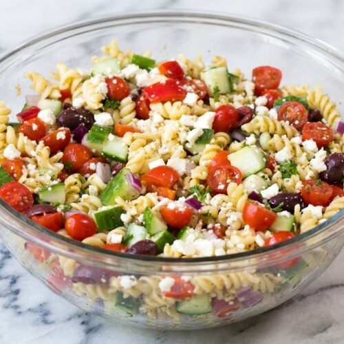 Traditional Greek Pasta Salad with feta-brine coating on al dente rotini in a minimalist ceramic bowl on white marble.
